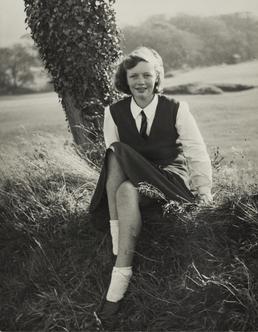 Kathleen Brew, seated on grass bank near tree…